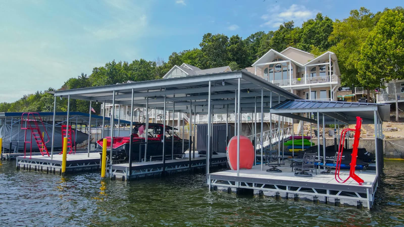 Two covered docks at Lake of the Ozarks with boats in the slips, a red water slide and a lounge deck, homes and trees on the hill behind