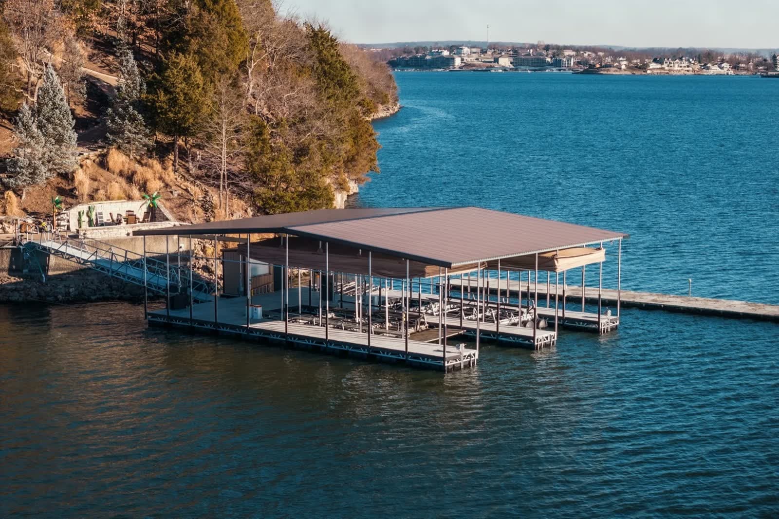 Covered floating dock with a brown metal roof on a rocky, wooded Lake of the Ozarks shoreline, trees bare in late fall